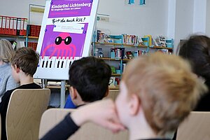 Kinder hören einem Vortrag zu, im Hintergrund ein Plakat der Kinderuni