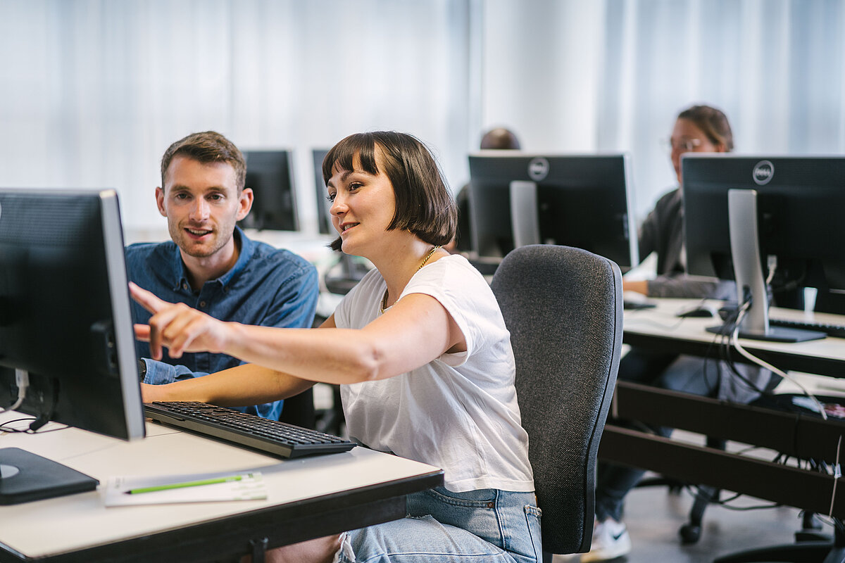 Informatik-Lehrraum mit Tischen und Computern. Im Vordergrund zeigt eine Studentin ihrem Sitznachbarn etwas auf dem Bildschirm.