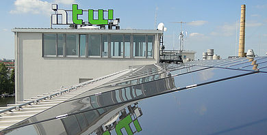 Solarzellen auf einem Dach auf dem Campus Wilhelminenhof, im Hintergrund das Hochschullogo